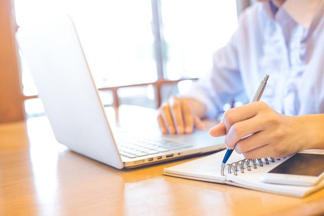 The hand of a businesswoman using a laptop and writing with a pen in Notepad.